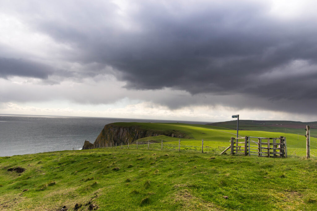 Stormy clouds seen from a green cliff side with a short piece of fence in the foreground.